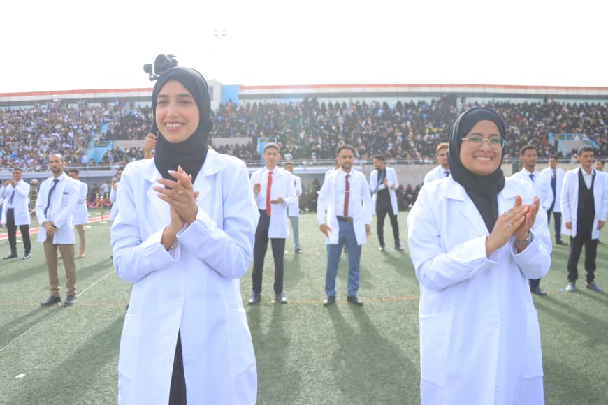 1003982394 - جامعة 21 سبتمبر Two women in white lab coats and hijabs clap on a football field during a graduation ceremony, with a crowd in the stands behind them.