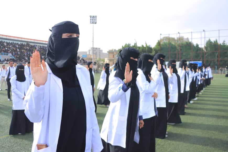 1003982397 - جامعة 21 سبتمبر Group of women in white lab coats and black hijabs standing in a line on a sports field, raising their right hands in a salute.