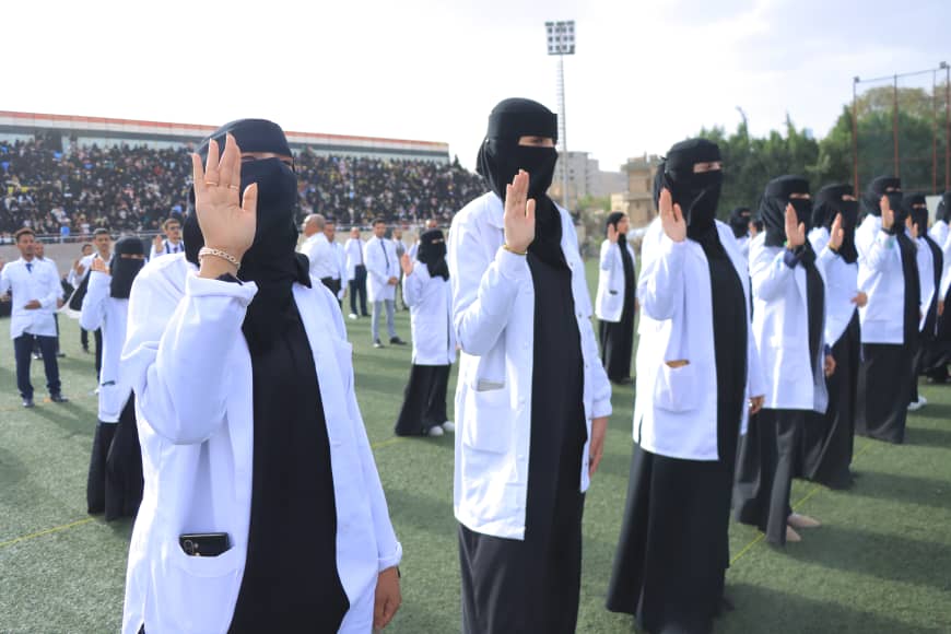1003982400 - جامعة 21 سبتمبر Women in white lab coats and black hijabs raise their right hands in a pledge during a ceremony at a stadium. Implementation: a formal oath among students or staff with a crowd watching in the background.