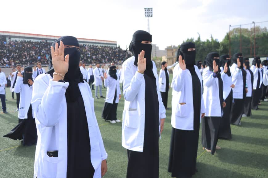 1003982405 - جامعة 21 سبتمبر Group of women in white lab coats and black niqabs raising their right hands in a ceremonial pledge on a football stadium field, with spectators in the stands.