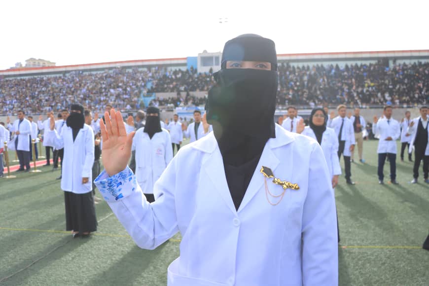 1003982408 - جامعة 21 سبتمبر Front-row woman in a white lab coat and black niqab raises her hand at a daytime outdoor ceremony on a stadium field, with a large crowd behind.