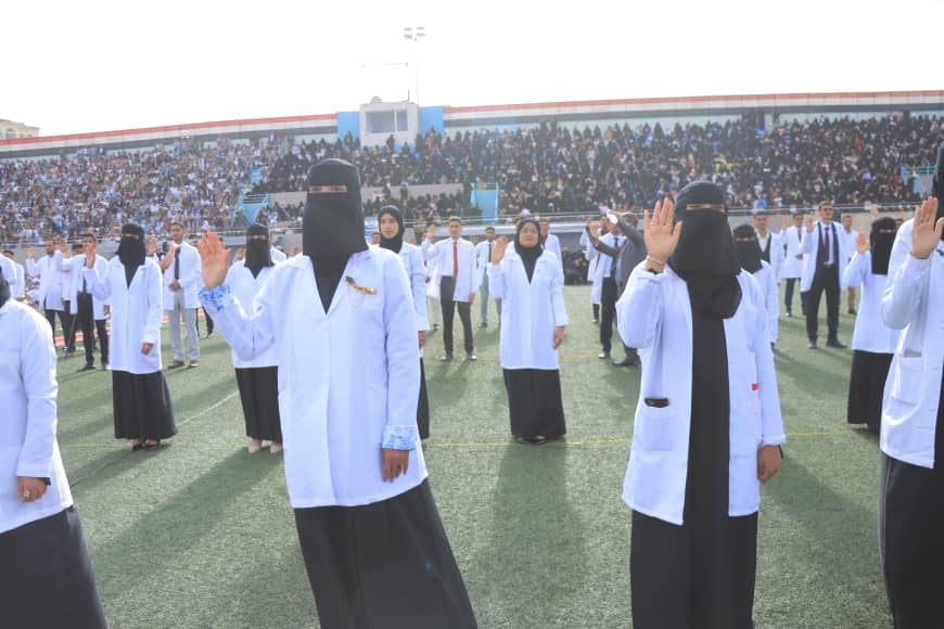 1003982411 - جامعة 21 سبتمبر Group of women in white lab coats and black head coverings saluting on a sports field during a ceremony, with a large crowd in the stands behind them.