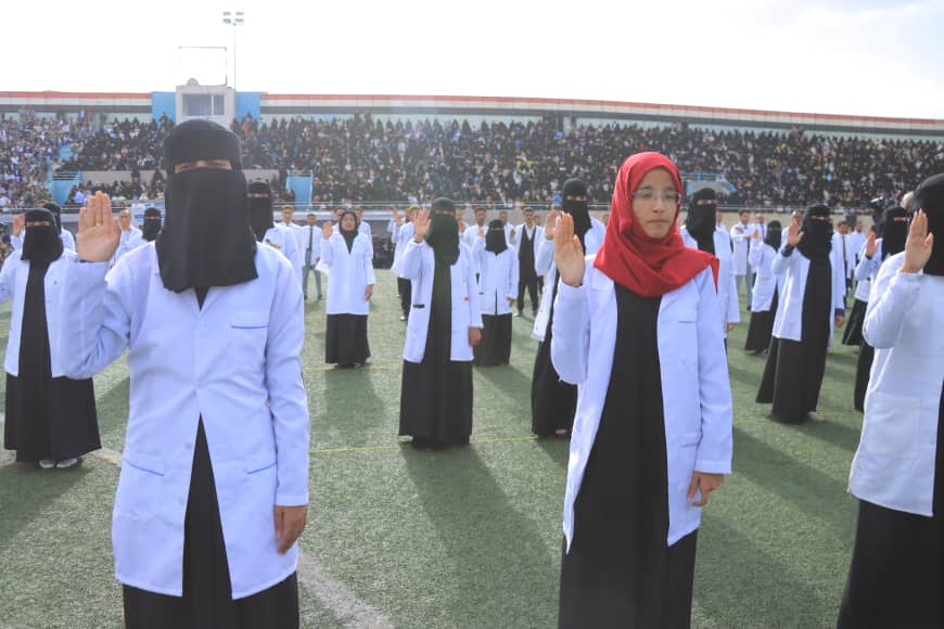 1003982414 - جامعة 21 سبتمبر Group of women in white lab coats raise their right hands in a ceremony on a sports field, with a stadium full of spectators in the background.