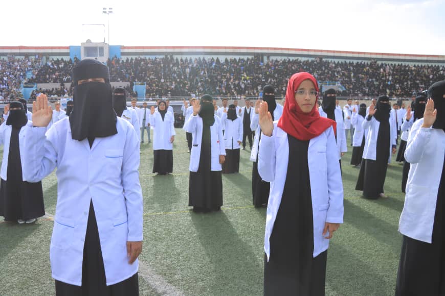 1003982417 - جامعة 21 سبتمبر Group of students in white lab coats over black dresses perform a right-hand salute on a field; a woman in a red hijab stands center with a stadium audience behind.