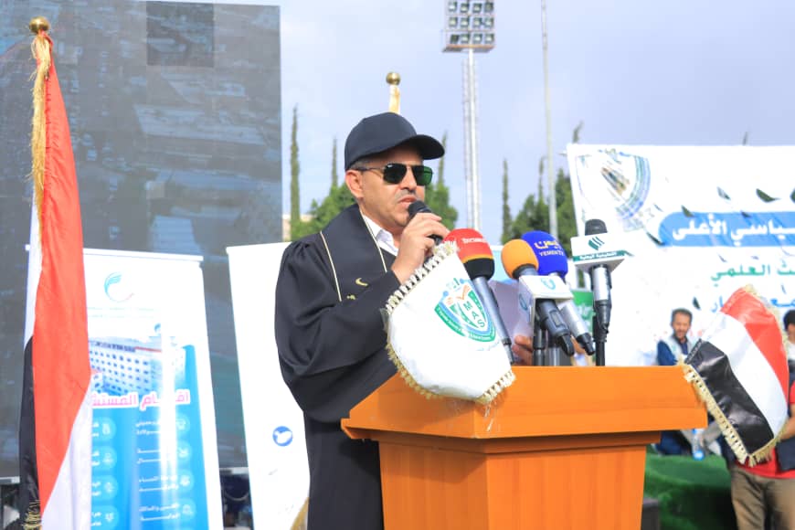 1003982420 - جامعة 21 سبتمبر Man in a cap and dark robe speaks into a microphone at a wooden podium, with multiple microphones and banners in the background at an outdoor event.