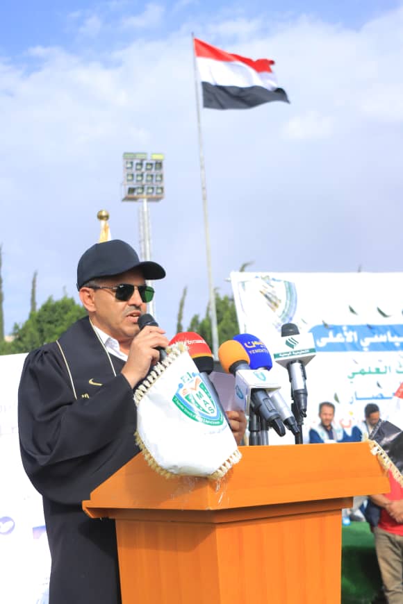 1003982423 - جامعة 21 سبتمبر Man in a black cap and sunglasses speaks at a wooden podium with multiple microphones, Egyptian flag in the background.