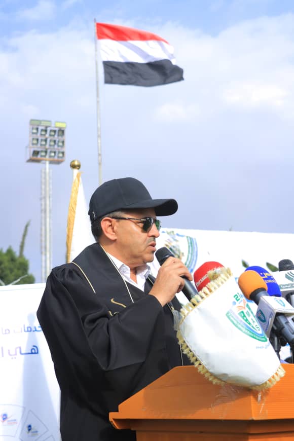 1003982426 - جامعة 21 سبتمبر A man in a black cap and sunglasses speaks into a microphone at a podium during a public event, with a red‑white‑black flag in the background.