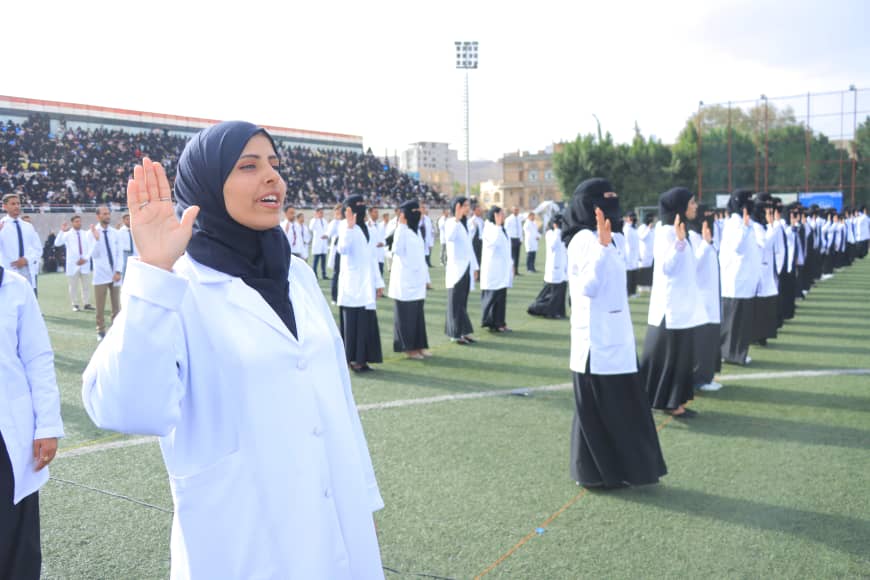1003982429 - جامعة 21 سبتمبر A woman in a hijab leading a lineup of students in white coats saluting on a sports field; audience in stands behind them.