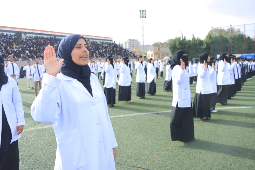 1003982432 - جامعة 21 سبتمبر Woman in a white lab coat and hijab raises her right hand as she leads a pledge on a sports field, with rows of clinicians behind her.