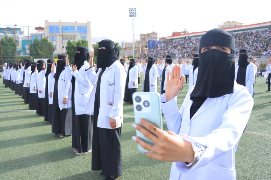 1003982435 - جامعة 21 سبتمبر Group of women in white coats and black niqabs standing in rows on a sports field during a ceremony, with a woman in the foreground taking a selfie using a blue phone.