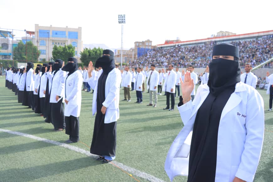 1003982438 - جامعة 21 سبتمبر Line of women doctors in white coats and black hijabs/niqabs standing in a line on a stadium field, raising a hand in a salute as spectators watch.