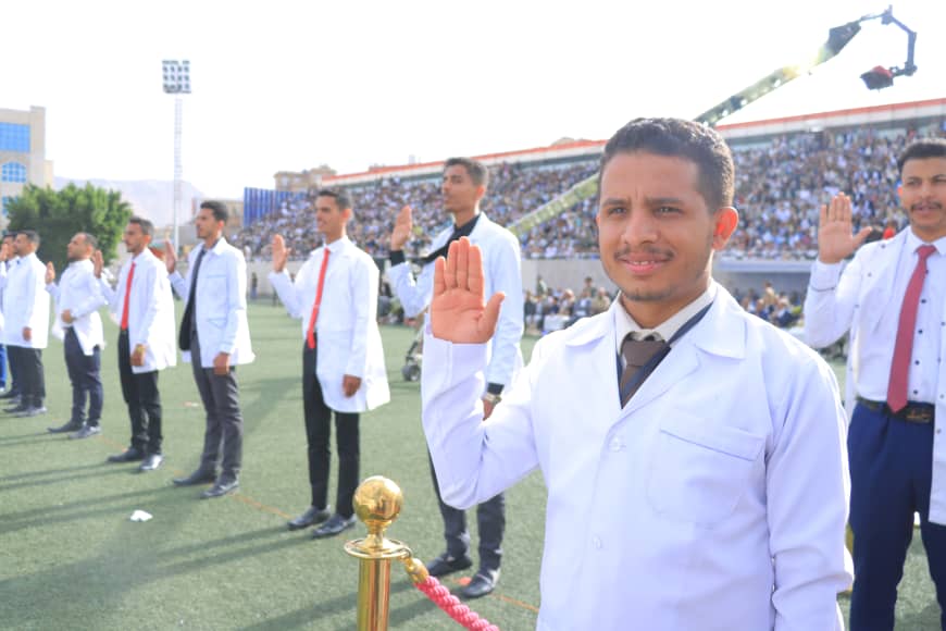 1003982441 - جامعة 21 سبتمبر A man in a white lab coat raises his right hand in a pledge on a sports field, with teammates and a stadium crowd behind him.