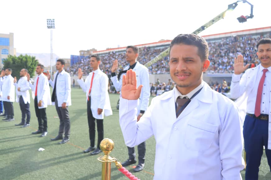 1003982444 - جامعة 21 سبتمبر Man in a white lab coat raises his right hand during a pledge on a sports field, with fellow graduates and a cheering stadium in the background.