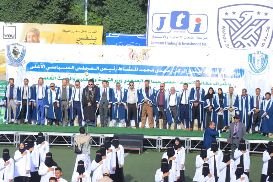 1003982447 - جامعة 21 سبتمبر Group of graduates in blue ceremonial robes on a stage at an outdoor ceremony, with a large banner and logos behind them.