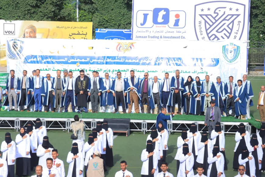 1003982450 - جامعة 21 سبتمبر Group of graduates in blue gowns on a stage at an outdoor ceremony, with banners in the background and a crowd below