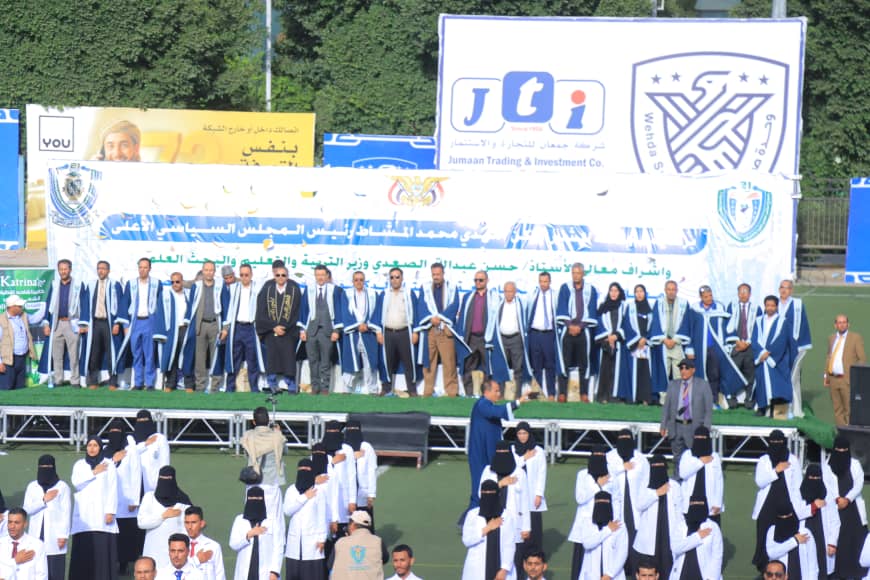 1003982453 - جامعة 21 سبتمبر Group of graduates in blue gowns standing on a stage during a ceremony with banners behind them.