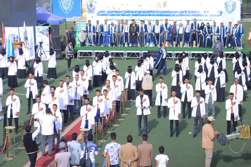 1003982456 - جامعة 21 سبتمبر A large group of graduates in white coats stand on a sports field with hands over their chests during a ceremony, while a blue-clad stage and banner loom in the background.