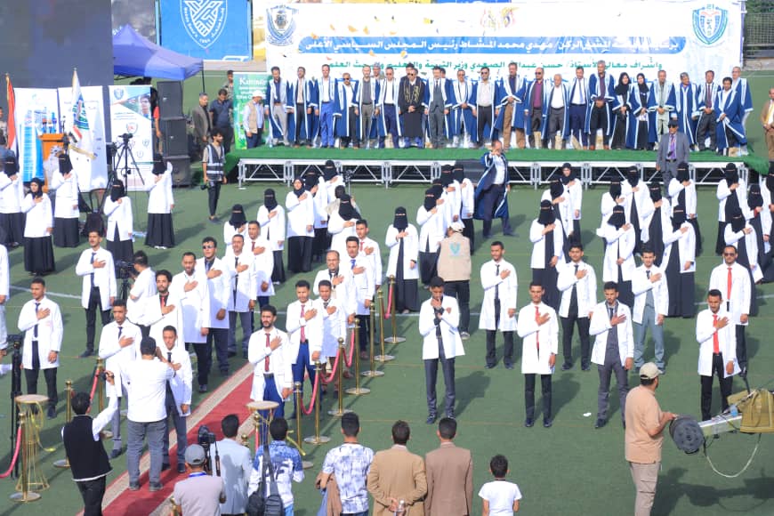 1003982459 - جامعة 21 سبتمبر Outdoor graduation ceremony with rows of graduates in white coats and hands over hearts, stage with blue-gowned faculty behind banners.