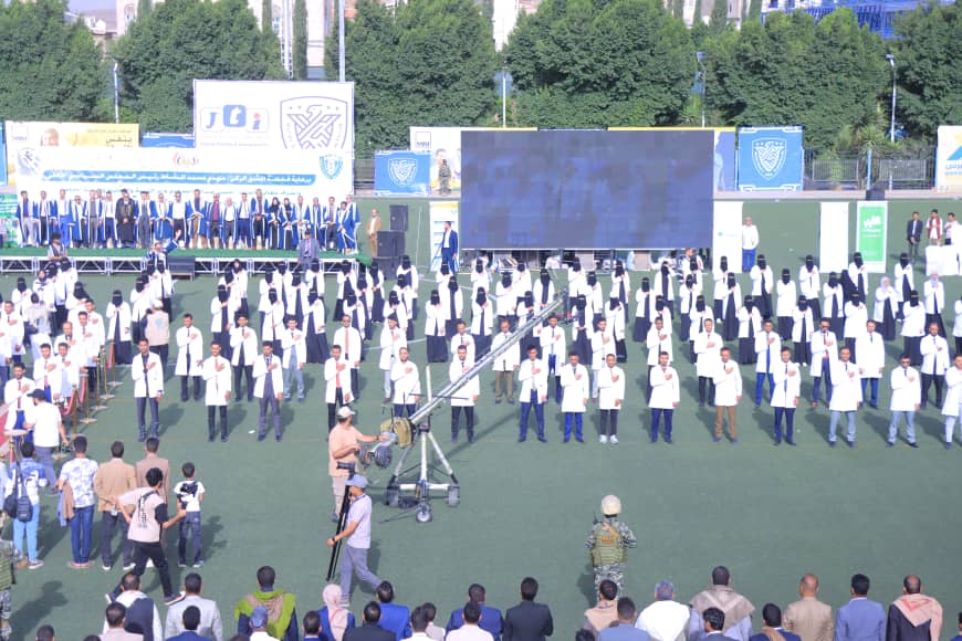 1003982462 - جامعة 21 سبتمبر Outdoor ceremony on a sports field with dozens of graduates in white coats standing in rows, a stage and large screen behind them, and a camerabot at center.