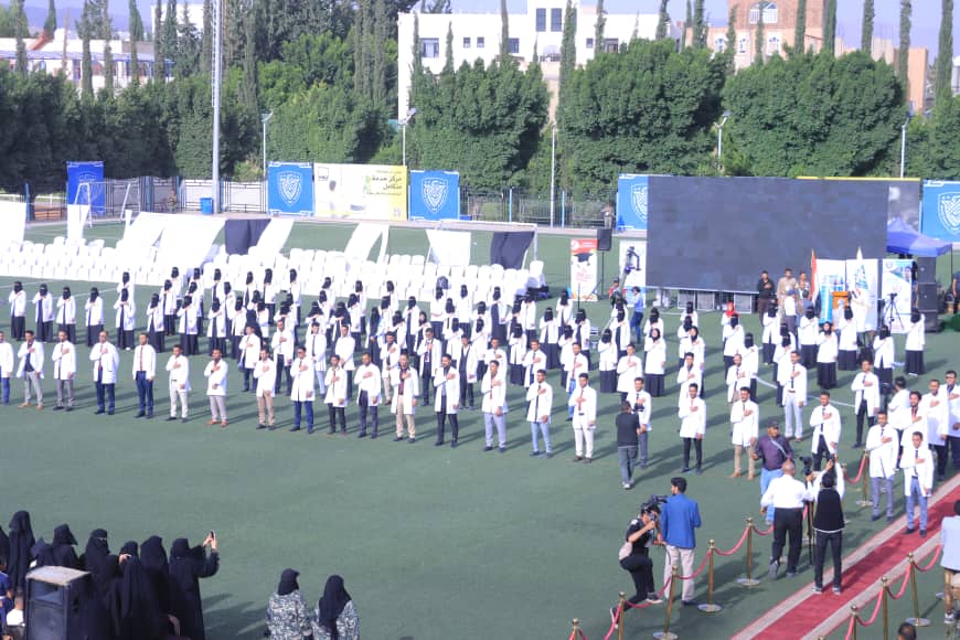 1003982465 - جامعة 21 سبتمبر Large outdoor ceremony with people in white coats standing in rows on a sports field near a stage and large screen.