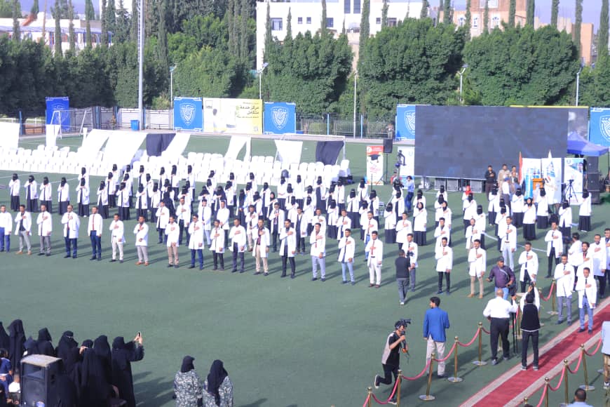 1003982468 - جامعة 21 سبتمبر Group of people in white coats standing in rows on a sports field during a ceremony, with a large screen and banners in the background.