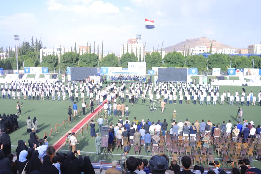 1003982471 - جامعة 21 سبتمبر Large outdoor ceremony on a sports field with students in white uniforms forming rows and an audience watching.