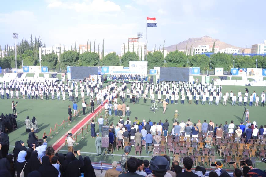 1003982472 - جامعة 21 سبتمبر Large outdoor ceremony on a football field with students lined up in neat rows around a red carpet; audience seated in front and a stage with screens in the background, flag flying above.