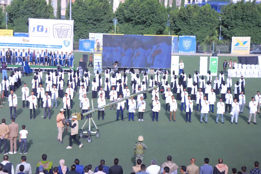 1003982473 - جامعة 21 سبتمبر Large group of people in white coats arranged on a sports field for a ceremony, with a big screen and camera crane in the center.
