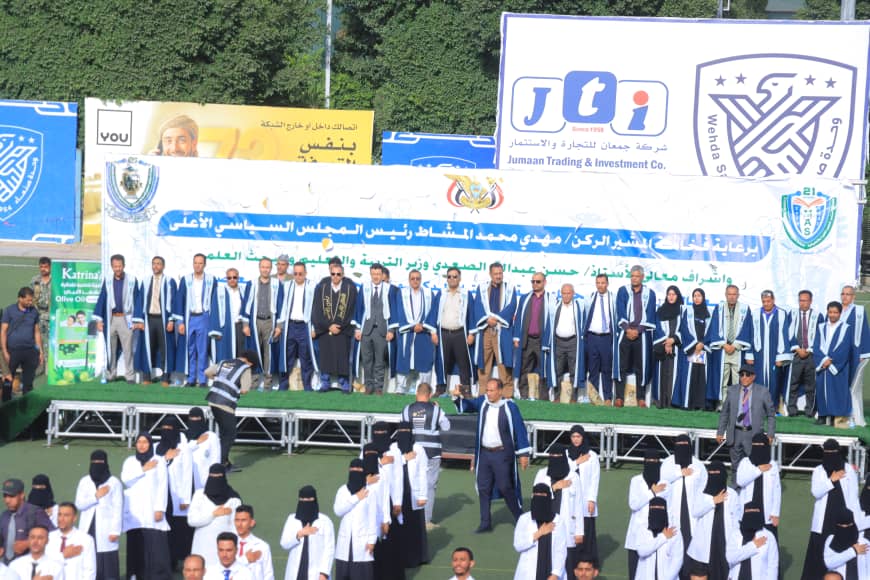 1003982474 - جامعة 21 سبتمبر Group of formally dressed people in blue robes on a stage during a ceremony, with large banners and signs in Arabic in the background.
