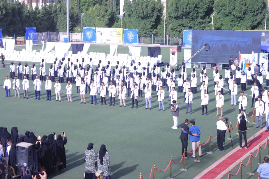 1003982475 - جامعة 21 سبتمبر Large group of people in white coats standing in rows on a sports field during a ceremonial event on a sunny day.