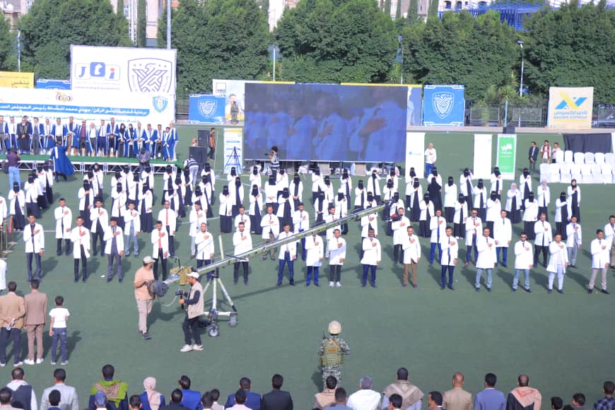 1003982476 - جامعة 21 سبتمبر Group of graduates in white coats standing on a sports field during a ceremony, with a large blue screen behind them and a camera crane in the foreground.