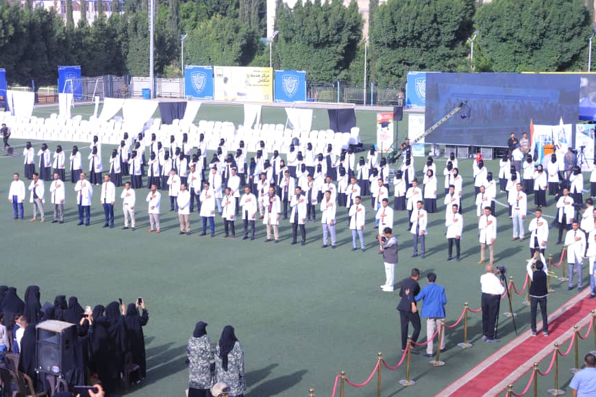 1003982477 - جامعة 21 سبتمبر Line of people in white coats standing in rows on a sports field during an outdoor ceremony, with banners and a stage in the background.