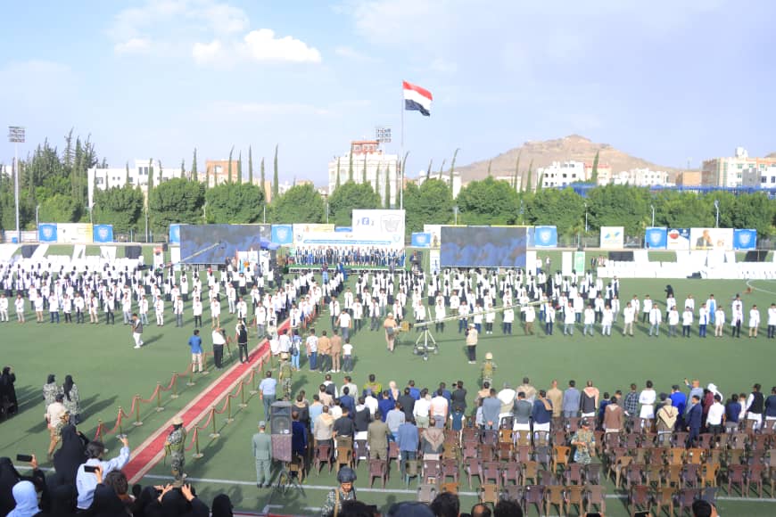 1003982478 - جامعة 21 سبتمبر Formal ceremony on a sports field with rows of people in white shirts facing a stage, Egyptian flag flying above and screening screens in the background.