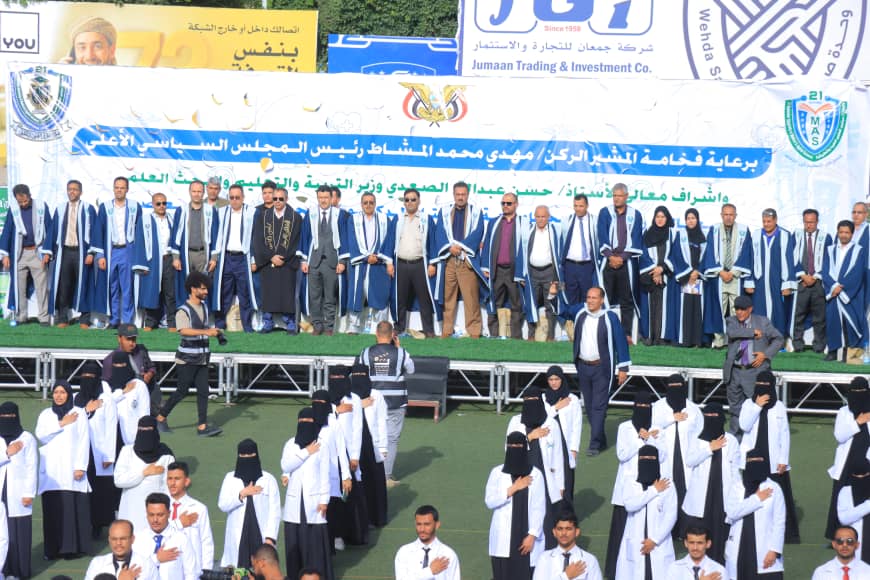 1003982479 - جامعة 21 سبتمبر Group of graduates in blue and white robes standing on stage during a formal ceremony, with an Arabic banner behind them on a sunny day Sympathetic crowd in audience and officials nearby.