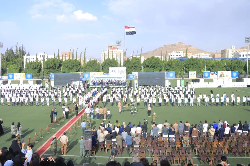 1003982498 - جامعة 21 سبتمبر Large outdoor ceremony on a football field with rows of people in white, facing a stage under a red‑white‑black flag above the venue.