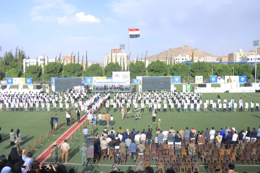 1003982502 - جامعة 21 سبتمبر Large stadium field ceremony with rows of uniformed participants facing a stage, audience in chairs, and a national flag flying above.