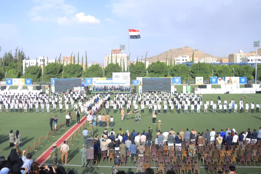 1003982505 - جامعة 21 سبتمبر Large outdoor ceremony on a sports field with rows of people in white uniforms forming geometric patterns, audience seated in front.