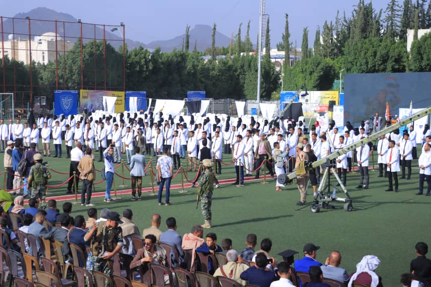 1003982508 - جامعة 21 سبتمبر Large outdoor ceremony with rows of people in white lab coats lined up behind a rope barrier on a sports field, nearby cannon on wheels and military personnel present, audience seated in front