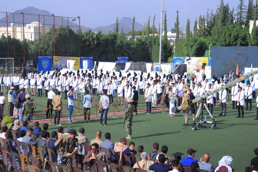 1003982511 - جامعة 21 سبتمبر Crowd watches a military demonstration on a sports field; rows of people in white coats stand behind ropes, with a mounted artillery piece nearby.