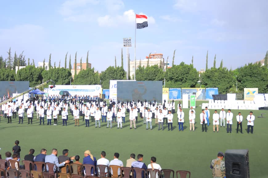 1003982514 - جامعة 21 سبتمبر Outdoor ceremony on a green field: students in white coats stand in rows, audience seated, with banners in the background.