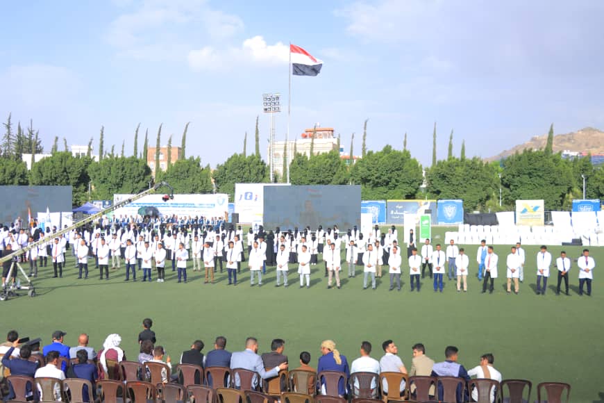 1003982517 - جامعة 21 سبتمبر Large group of people in white coats lined up on a green field during an outdoor ceremony, with spectators seated in front and banners in the background.