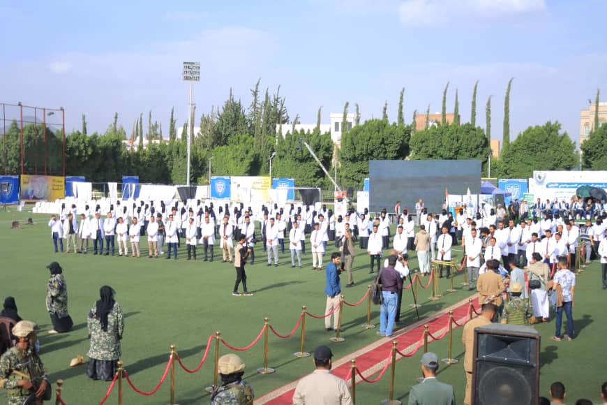 1003982520 - جامعة 21 سبتمبر Line of people in white uniforms stand in a long row on a grassy field during a formal ceremony, cordoned off by red ropes.