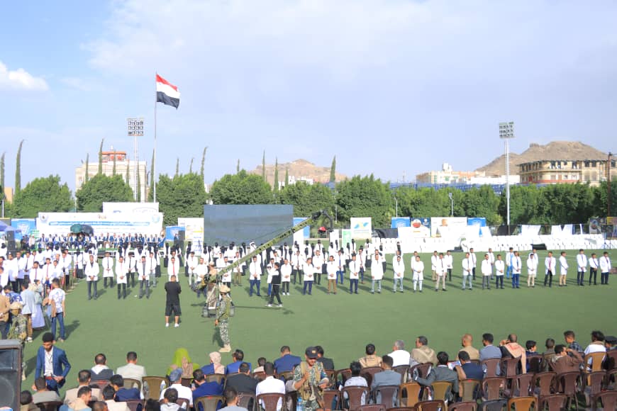 1003982523 - جامعة 21 سبتمبر Large outdoor ceremony on a grassy field with rows of people in white shirts, flag in the background, and an audience seated in the foreground.