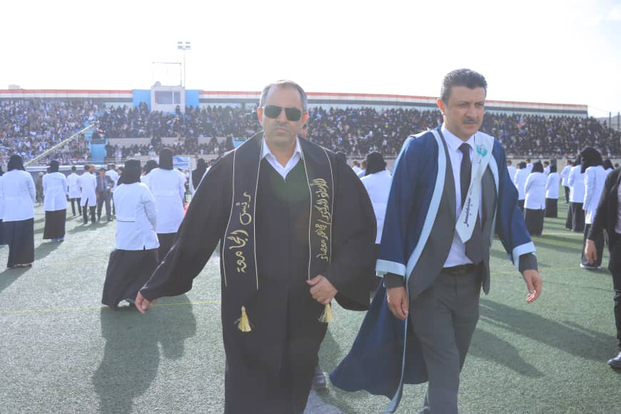 1003982526 - جامعة 21 سبتمبر Two men in academic gowns walk across a stadium field during a graduation ceremony as a large crowd watches from the stands in the background.