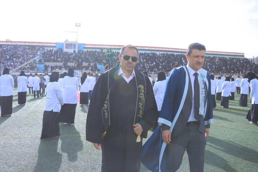 1003982529 - جامعة 21 سبتمبر Two men in graduation gowns stand on a sports field with a large audience in the stands behind them.