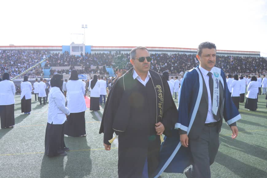 1003982532 - جامعة 21 سبتمبر Two men in graduation robes walk across a sports field as a large crowd watches from the stands behind them.