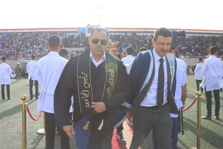 1003982535 - جامعة 21 سبتمبر Two men walk onto a stadium field for a ceremony; one wears a black ceremonial gown with gold embroidery and sunglasses, the other a suit with a blue strap.
