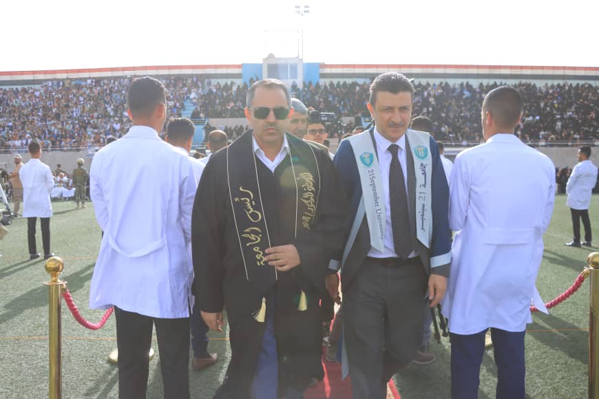 1003982538 - جامعة 21 سبتمبر Two men in ceremonial robes walk toward the camera on a red carpet at a crowded stadium with spectators in the stands behind them.