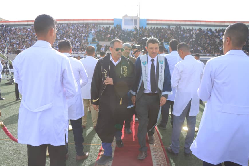 1003982541 - جامعة 21 سبتمبر Ceremonial procession on a red carpet in a stadium, with two men in robes flanked by staff in white coats and a large crowd watching.