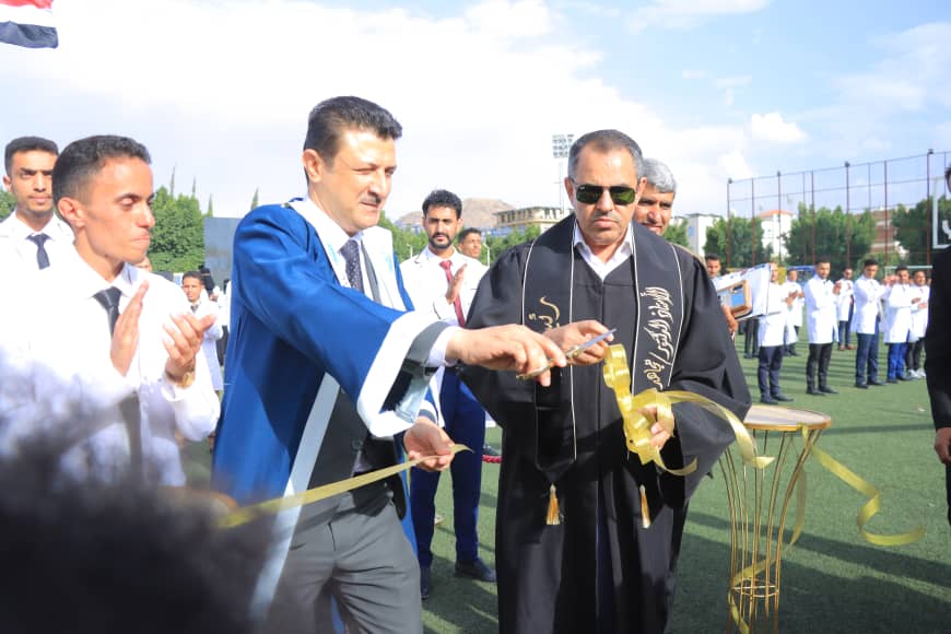 1003982544 - جامعة 21 سبتمبر Official in a blue gown cuts a ribbon with a man in black robes at a ceremonial opening on a sports field, with spectators in the background.